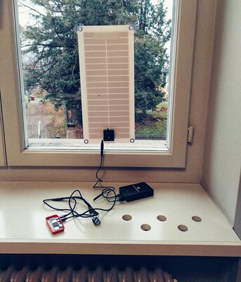 A solar panel attached to a window, connected to a battery. The battery is connected to a small red plastic casing, containing a Raspberry Pi